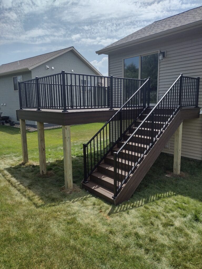 Elevated wooden deck with black metal railing and stairs attached to a house.