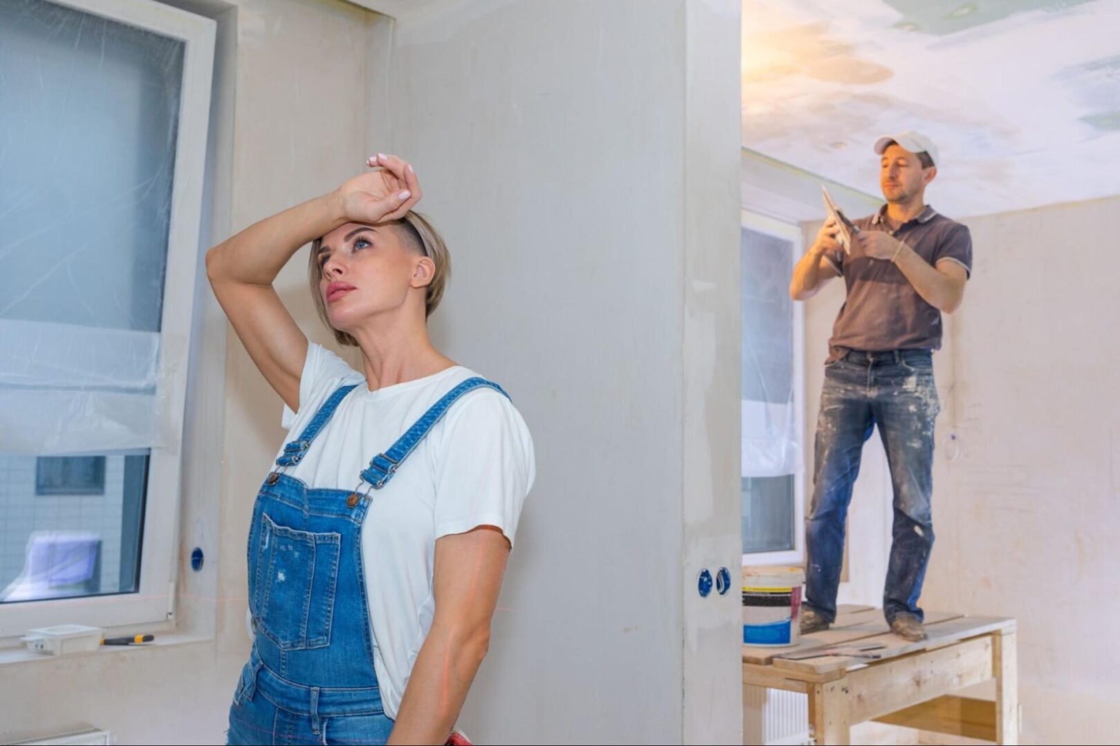 A man plasters walls and ceilings during house remodeling while a woman in denim overalls leans against a wall, appearing thoughtful and contemplative.