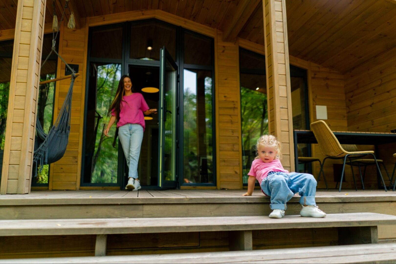 A smiling, cute little girl in stylish clothes sits on the terrace of a wooden house as her mom steps out through a modern patio door.