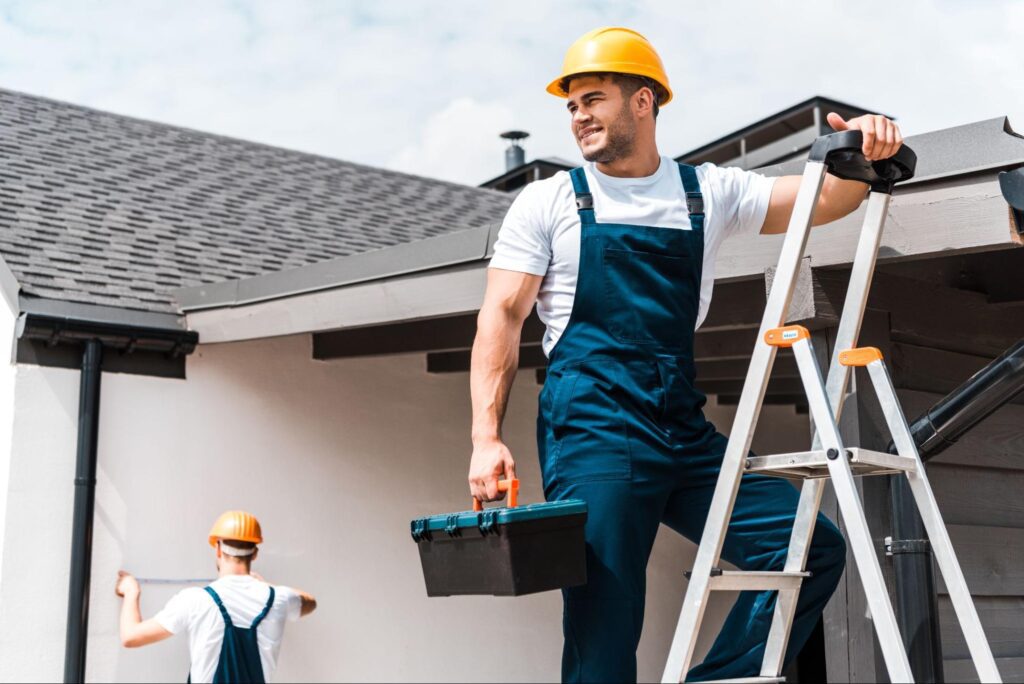 Man in a hard hat and holding a toolbox while using a ladder next to a roof.