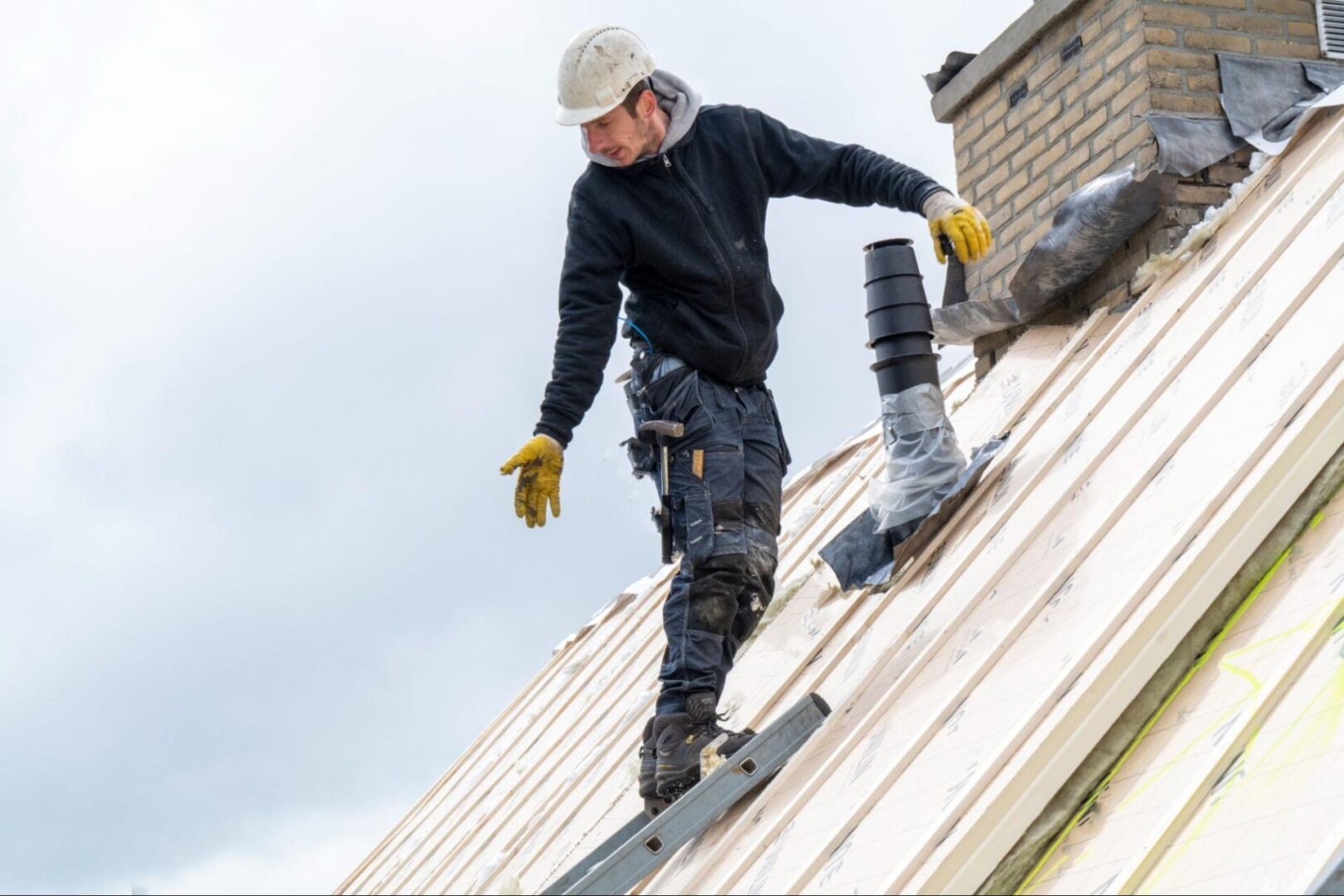 A roofer wearing a hard hat stands on a ladder, checking how long the roof lasts.