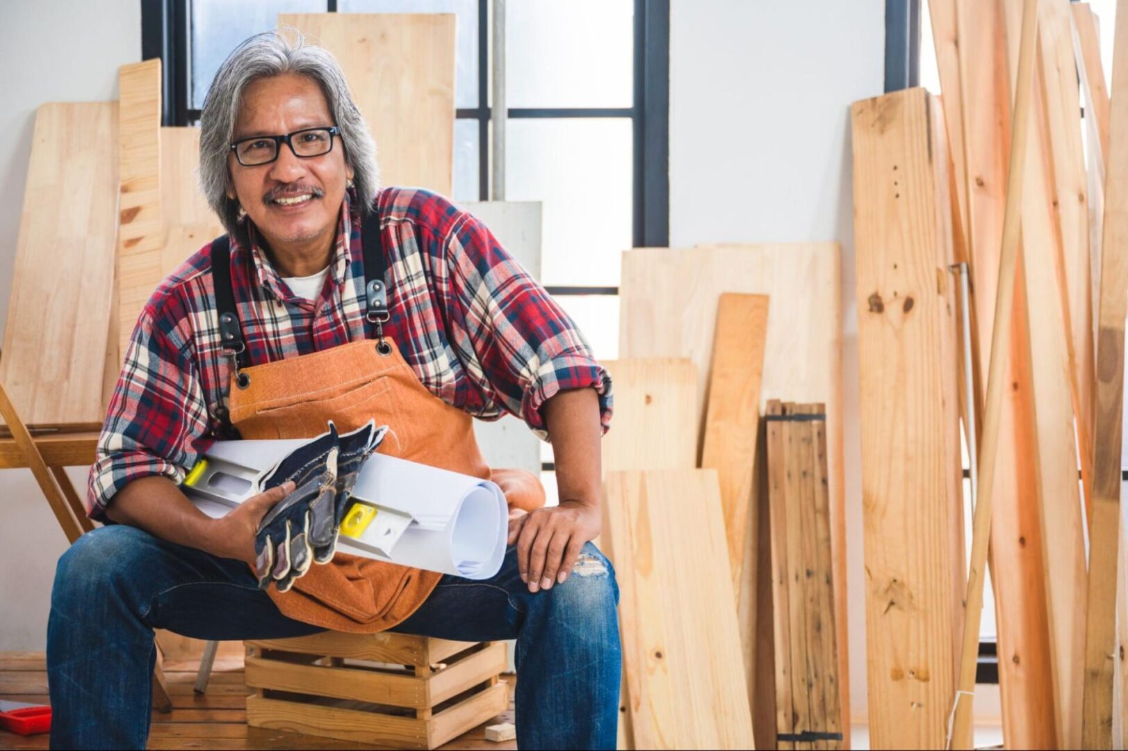 Man in an apron and holding gloves next to planks of wood. 