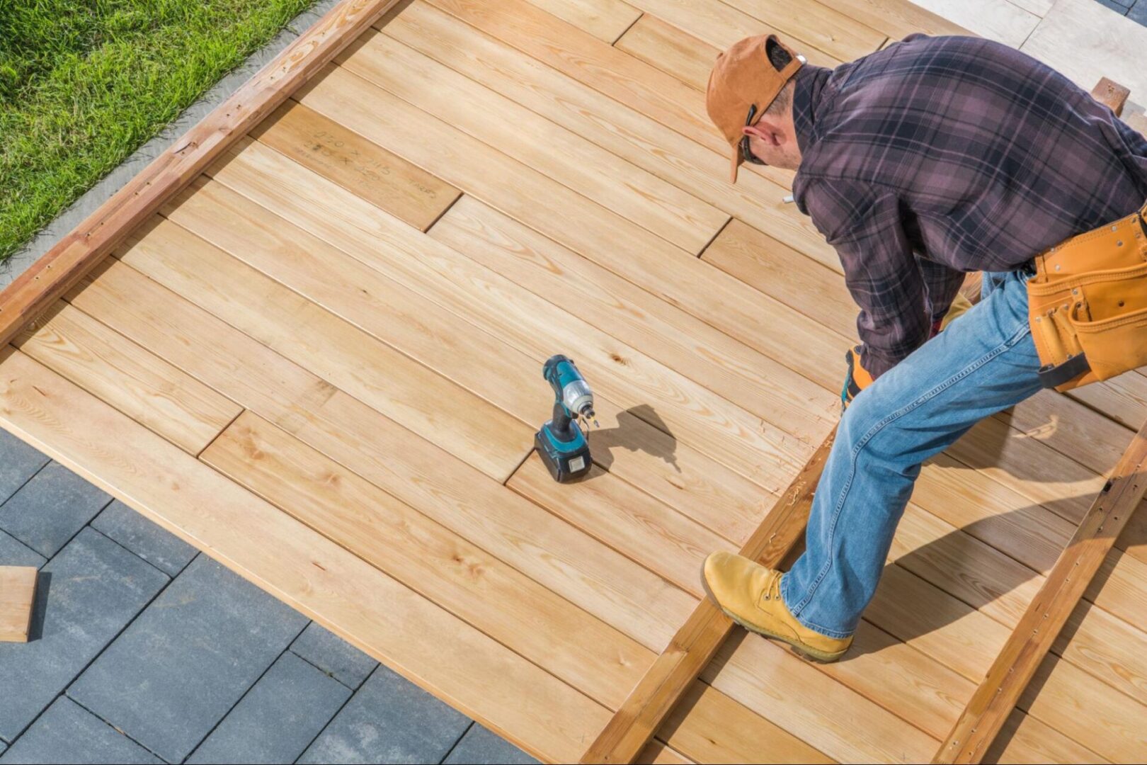 A carpenter arranges long wooden planks on top of wood tiles for a deck, with a drill nearby.