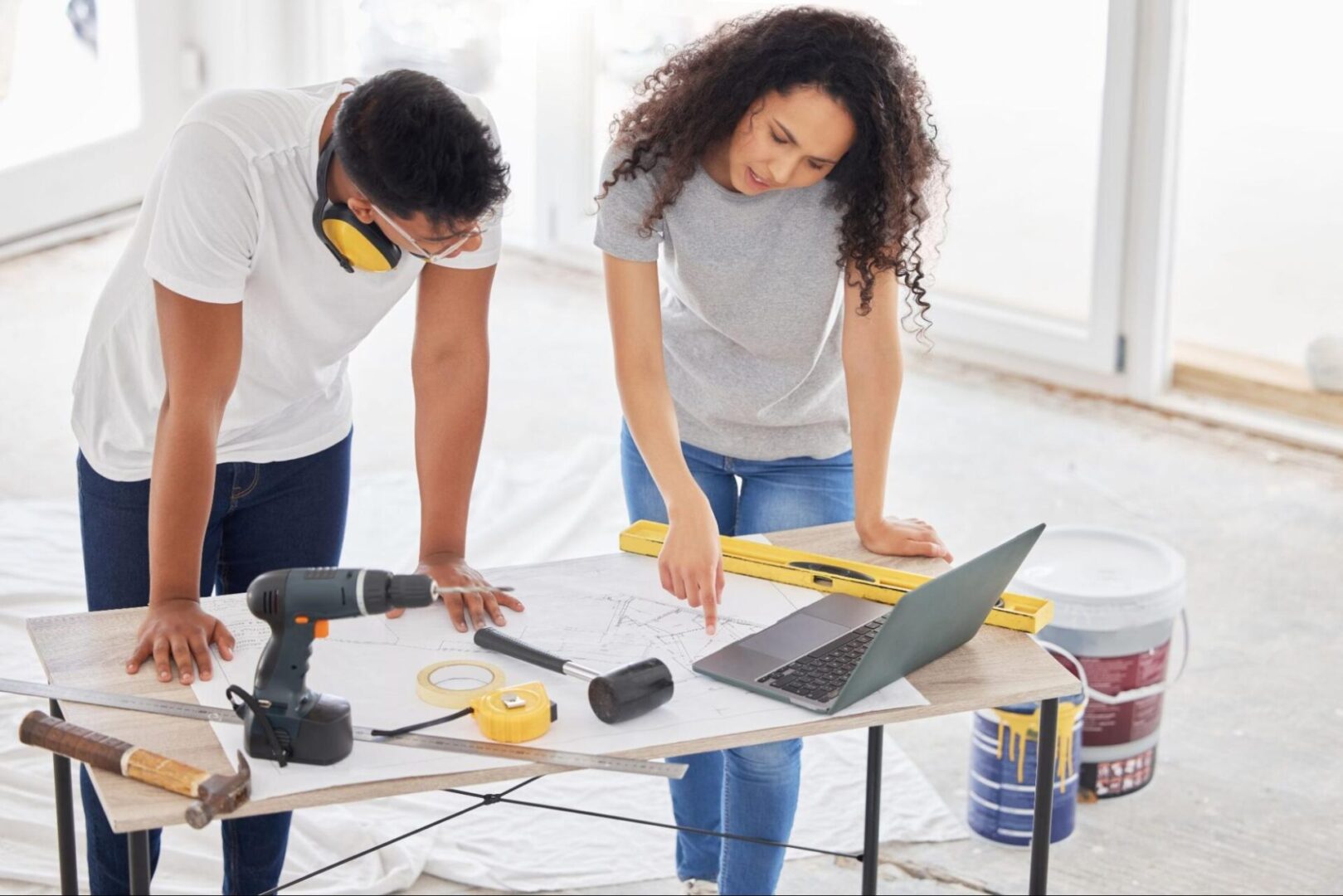 A couple reviews house remodeling plans on a laptop and orders materials online. Tools rest on the table, with a paint bucket beside a white protective floor covering nearby.