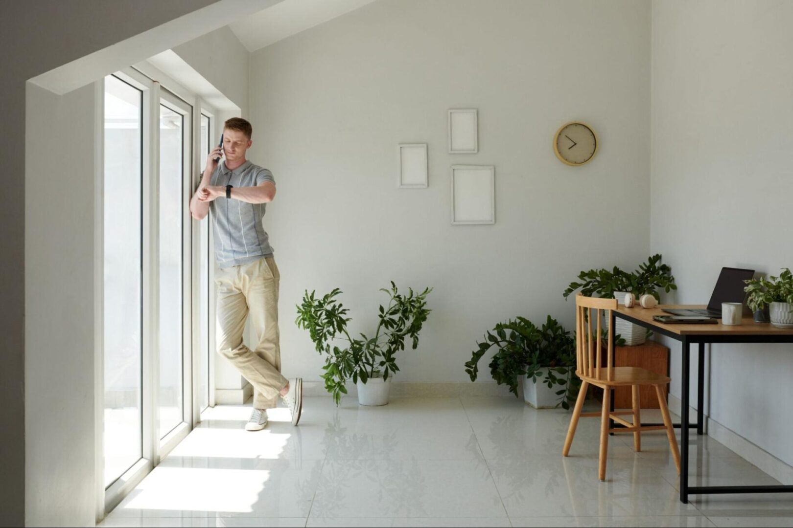 Natural light coming in through glass doors next to a man looking at his watch next to plants and a desk. 