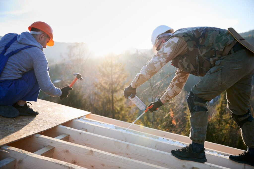 Two roofers wearing hard hats repair the roof to improve insulation.