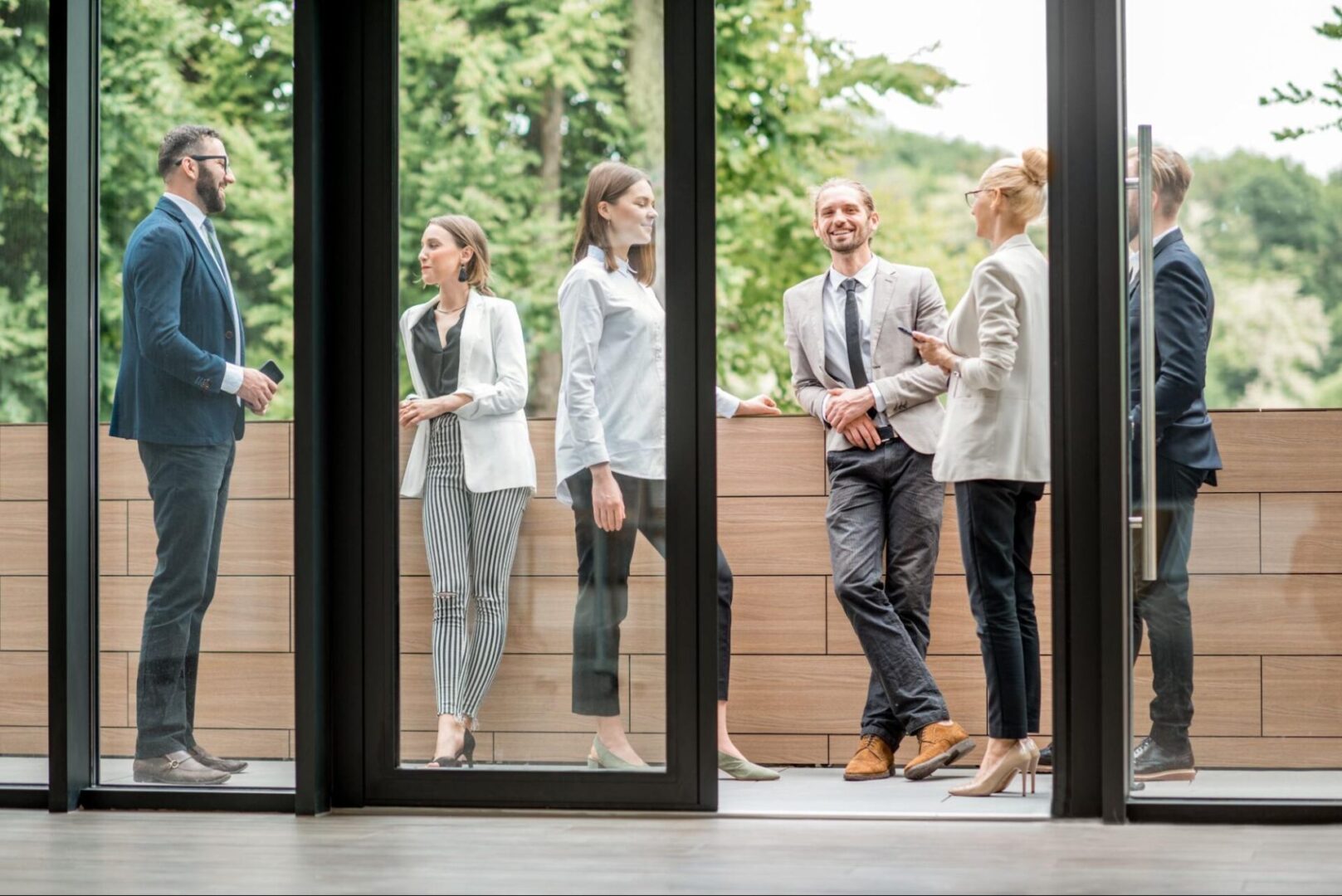 Colleagues standing near a black-framed glass door enjoy a break on an office balcony, 