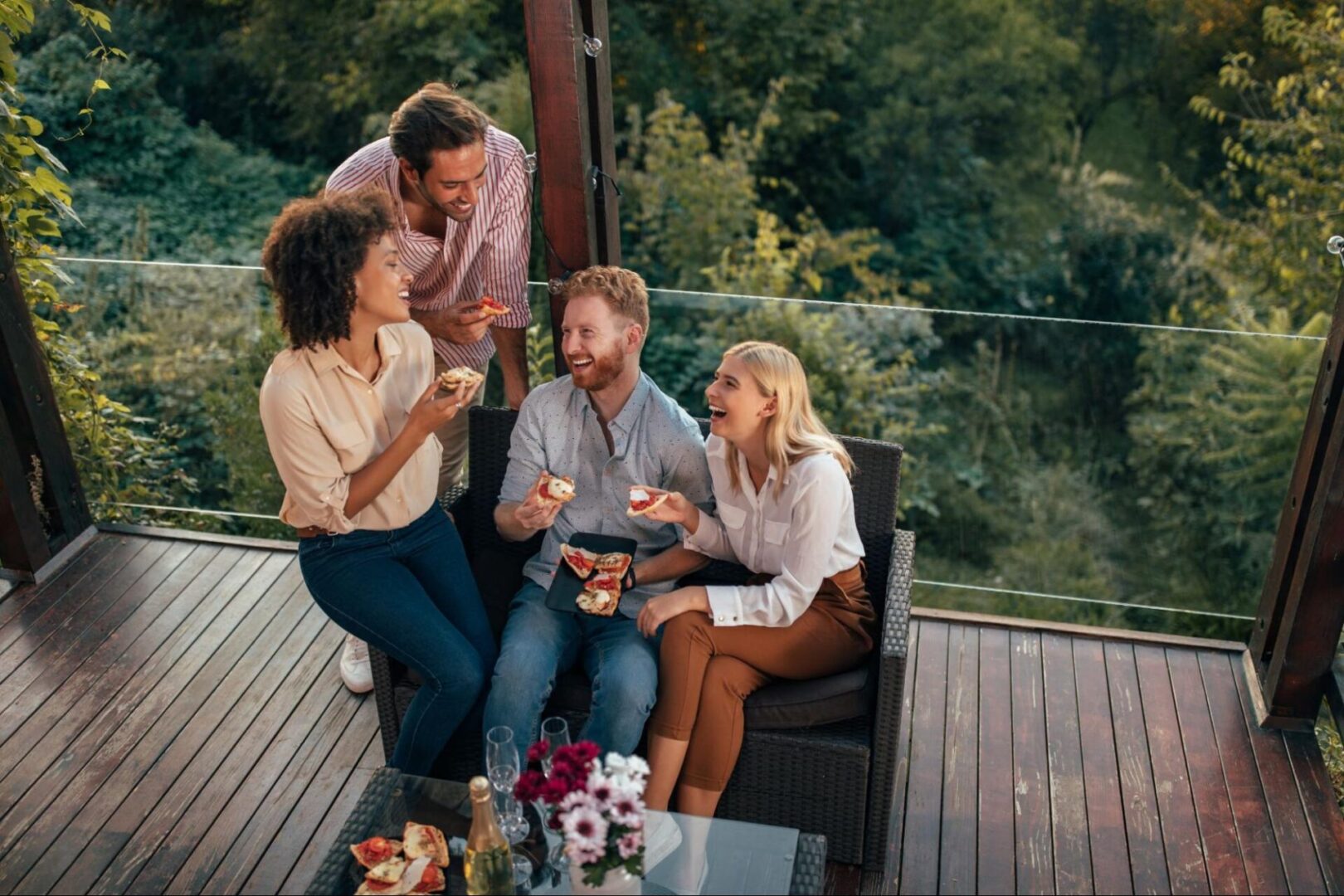 Friends eating pizza while standing and sitting on a wooden deck with a glass fence. 