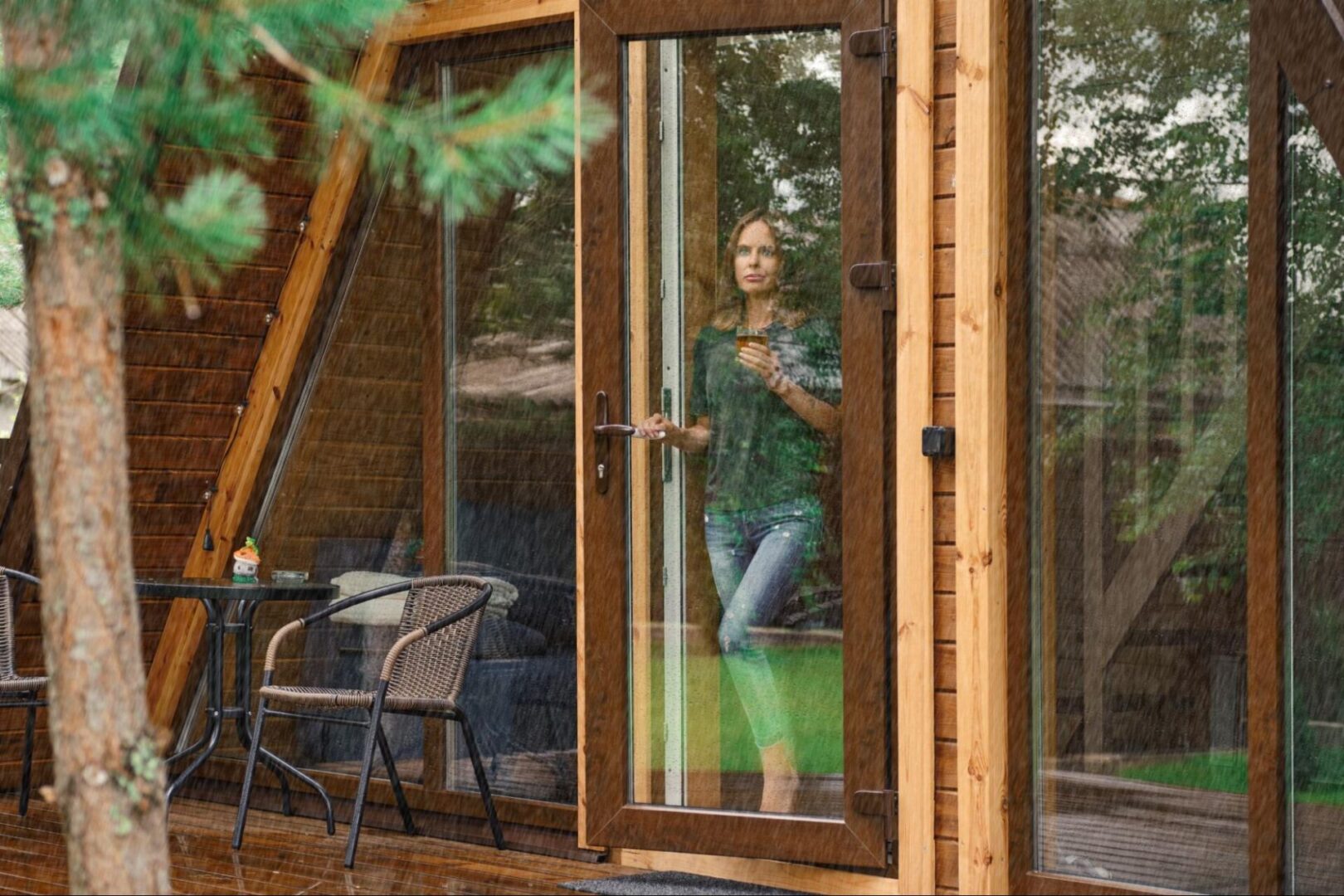 A young woman holding a cup of herbal tea stands by a forest house patio door, gazing thoughtfully at the rain falling outside.